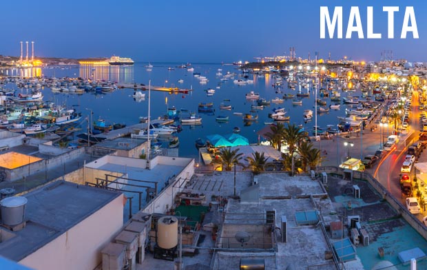 The island of Malta at dusk showing the soft blue lights of the harbour and the orange and yellow lights of the city